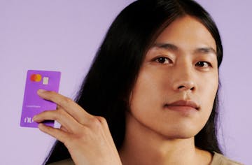 Young man on a neutral background looking at the camera and holding a Nubank card next to his face