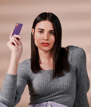 straight-haired woman wearing a grey sweater while looking at the camera and holding Nubank's business account card
