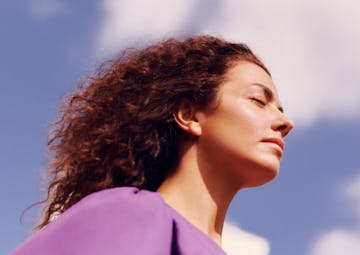 Mulher com cabelos cacheados soltos, olhos fechados, com o céu ao fundo