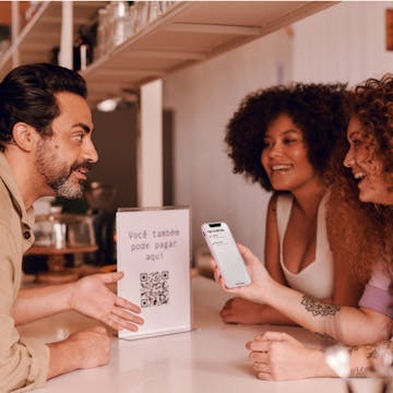 Duas mulheres apoiadas em um balcão comercial negociando pagamento por QR code com o vendedor.