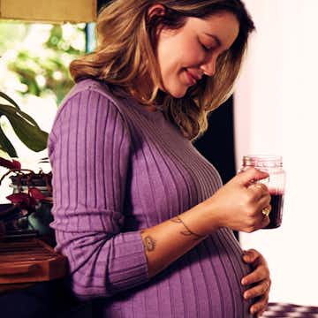 Image of a pregnant woman, wearing a purple blouse and holding a mug with juice.