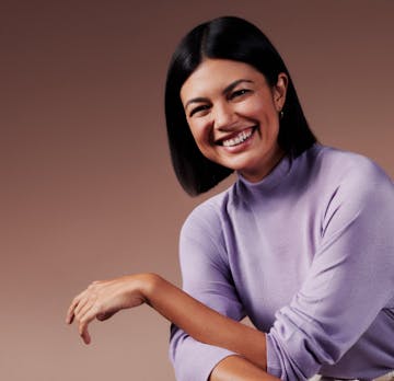 Mulher branca com cabelos curtos, liso e preto está sorrindo e vestindo uma blusa roxa.