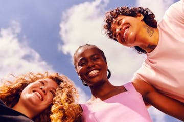 Três pessoas na imagem. À esquerda rosto de uma mulher de pele branca e cabelos loiros cacheados. Ao centro mulher de pele negra e cabelos curtos trançados, vestindo camiseta rosa, À direita rosto de homem de pele avermelhada com cabelos cacheados. Todos estão sorrindo com o céu azul com nuvens ao fundo.