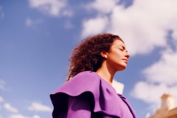 White-skinned woman, curly hair reddened back by the wind, wearing a purple blouse. She has her eyes closed. In the background blue sky and clouds.
