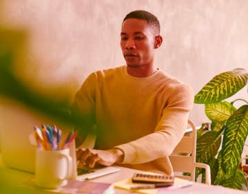 Black-skinned man wearing a long-sleeved beige shirt. He is being on a chair and typing on his laptop.