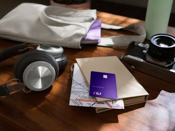 Foto da vista lateral de uma mesa de madeira, na qual estão dispostos, a frente, uma caixinha de som, uma caderneta com um mapa dentro e um envelope acima. Acima do envelope, também, tem-se o cartão Nubank. Ao fundo, é possível identificar uma ecobag com um caderno roxo em seu interior.