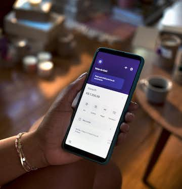 mulher em um ambiente de sala, segurando celular com a mão esquerda e utilizando o app Nubank