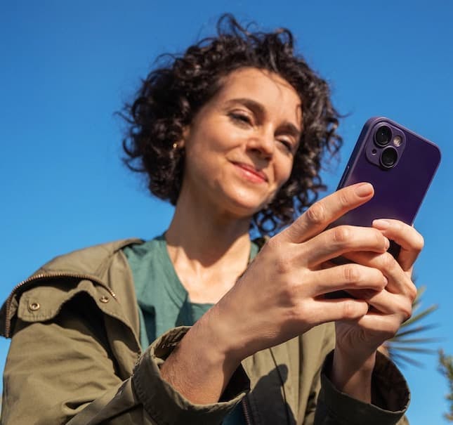 Foto mostra uma mulher branca, com cabelos curtos, escuros e ondulados, ao ar livre, segurando um celular com as duas mãos e olhando pra ele. O céu está azul. Ela veste camiseta verde e uma blusa verde oliva.