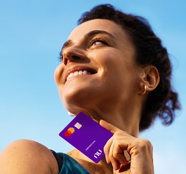 Foto mostra uma mulher branca, segurando um cartão roxinho do Nubank, ao ar livre. O céu está azul.