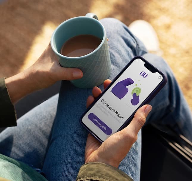 Foto destaca a mão de uma pessoa segurando um celular, com a tela para o app do Nubank, e com a outra mão segurando uma caneca com café.