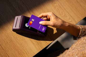 Foto de uma máquina de cartão em cima de uma mesa de madeira. Uma mão feminina está aproximando o cartão Nubank da máquina, representando a função contactless de pagamento.