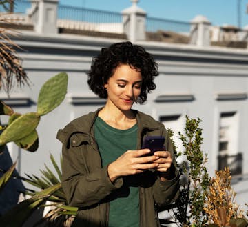 Retrato frontal de uma mulher branca, com cabelos cacheados acima dos ombros, com uma feição sorridente, segurando com as duas mãos um smartphone, o qual ela está olhando. Ao fundo, tem-se um muro branco e também algumas folhagens de plantas.