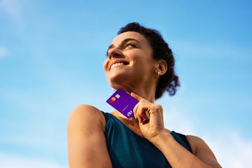 Retrato de uma mulher com cabelos cacheados curtos, com o rosto virado, sorrindo e olhando para o horizonte. A mulher veste uma regata verde petróleo. Em sua mão esquerda ela segura o cartão Nubank. No fundo, tem-se um céu azul com poucas nuvens.