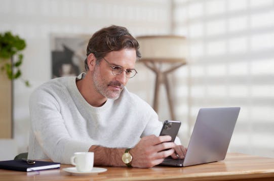 Homem branco sentado em uma mesa de trabalho com o computador aberto, enquanto olha o seu celular