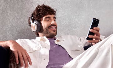Homem branco sentado de forma despojada no sofá, ele está de fone de ouvido enquanto usa o seu celular