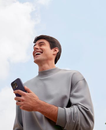 Homem branco com seu celular na mão, ele está sorrindo olhando para o céu.