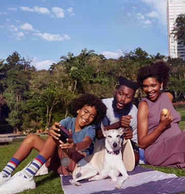 3 pessoas pretas sentadas na grama, sendo um casal e seu filho menor e um cachorro branco e bege.