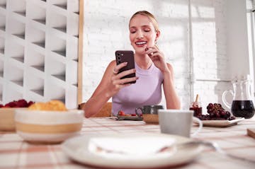 Foto de mulher branca sorrindo, segurando um celular, enquanto está lanchando em um mesa com alguns alimentos.